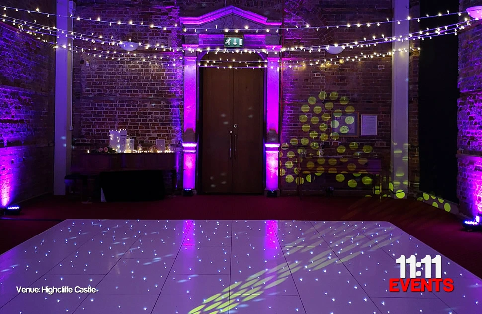 A white starlit dance floor installed at Highcliffe castle with a fairy light canopy from wall to wall and bright pink, magenta mood lights highlighting the pillars either side of a grand wooden door