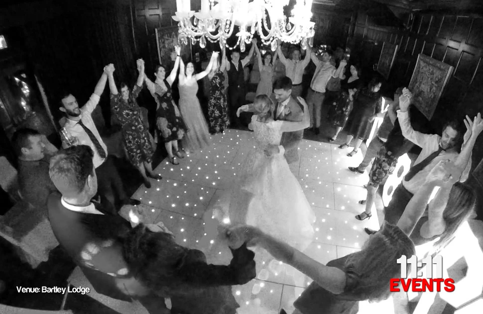 Black and white photo of a first dance at Bartley lodge taken from height looking down on the newly married couple dancing on a white starlit LED dance floor surrounded by guests with their hands raised and joined together
