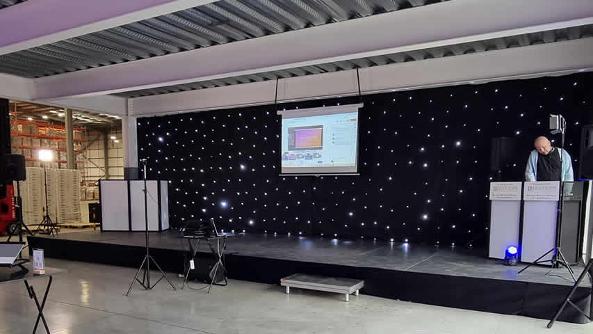AV for Corporate product launch with black stage, speakers, wireless microphones, DJ, mood uplighting and starcloth