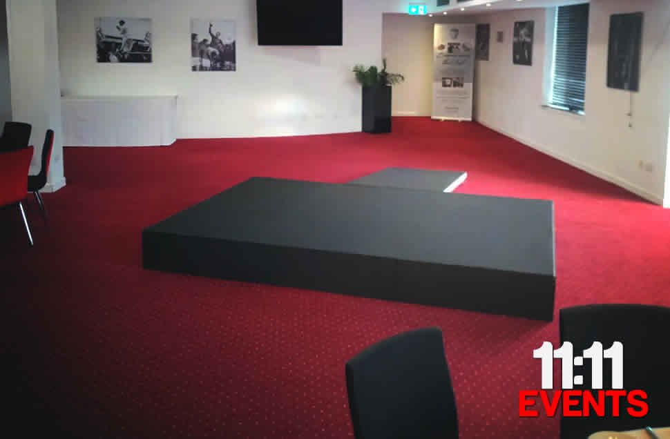A black carpeted stage with black valance skirt and black wheelchair ramp for a presentation. Placed on red carpet with white walls in Bournemouth