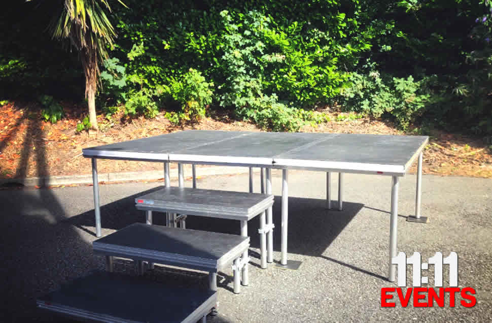A black topped stagedex stage outside on tarmac with 3 steps leading up to a 3x2m 1m high photoshoot platform with greenary in the backdrop. Bournemouth.